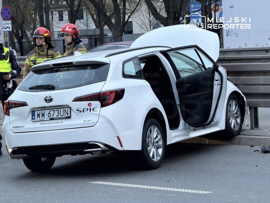 Biały samochód osobowy stoi na krawężniku z otwartymi drzwiami, obok trzech strażaków w hełmach.