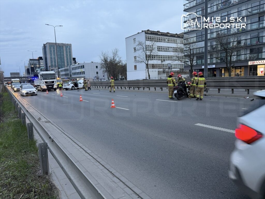Grupa strażaków udziela pomocy poszkodowanemu motocykliście na ruchliwej drodze, otoczeni przez pojazdy i znaki ostrzegawcze.