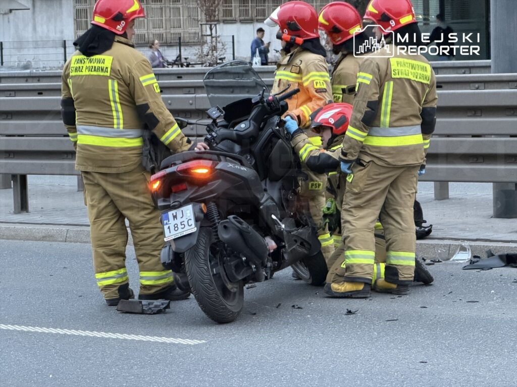 Grupa strażaków w mundurach z odblaskowymi elementami udziela pomocy poszkodowanemu na miejscu wypadku motocyklowego.