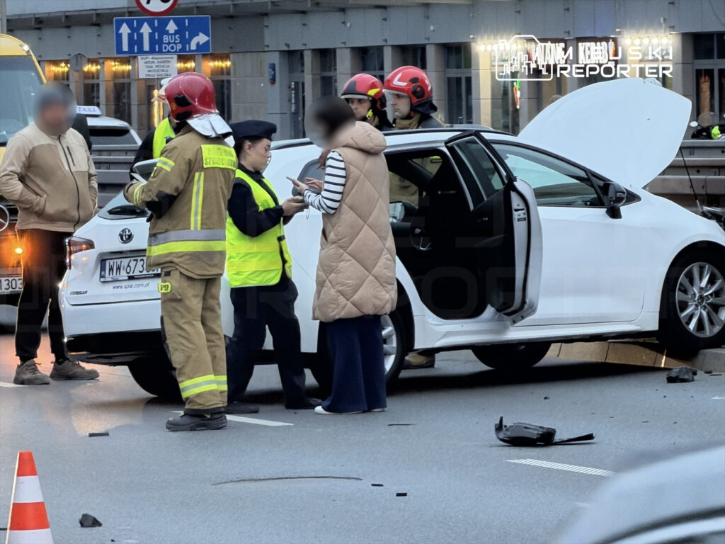 Grupa ratowników i policjantów rozmawia z kierowcą białego samochodu osobowego na drodze po wypadku.