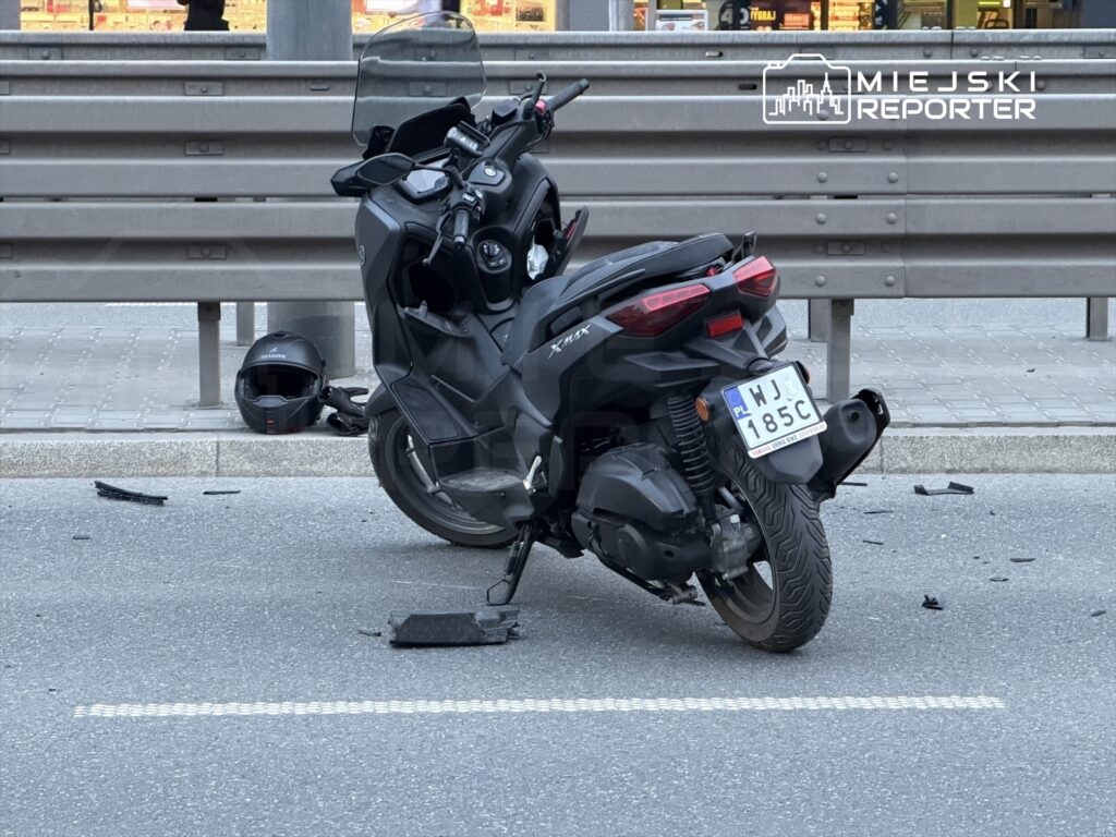 Zniszczony skuter leży na jezdni, obok niego kask oraz fragmenty plastikowe. W tle widoczny jest ruch uliczny.