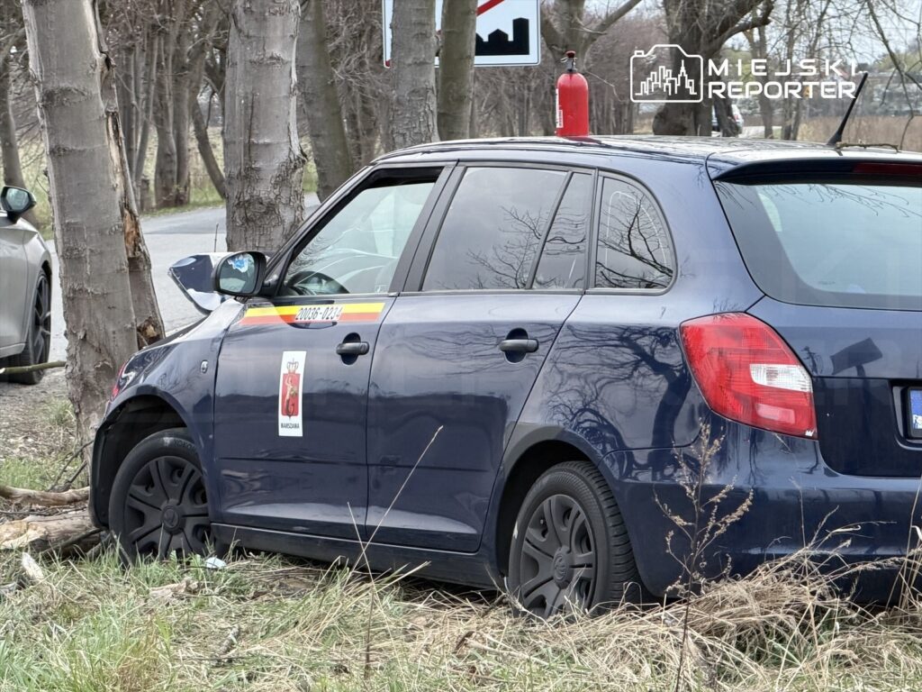 Nieoznakowany samochód osobowy z uszkodzoną lewą stroną stoi przy drodze, obok drzew i znaku drogowym.
