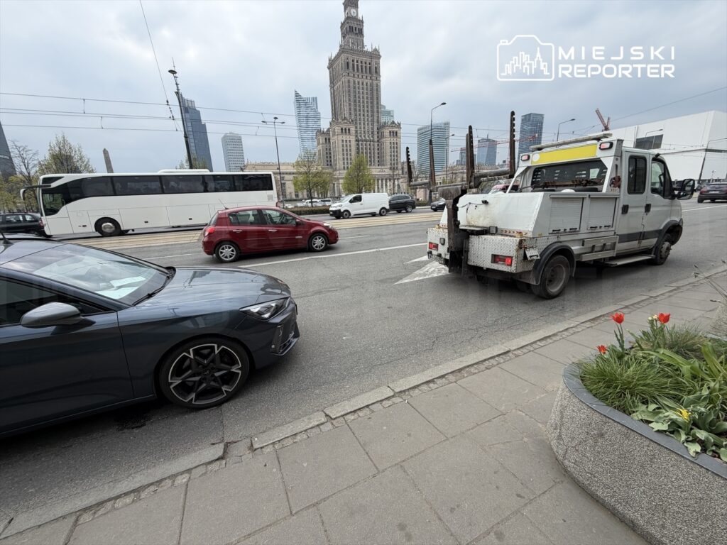 Czarny samochód osobowy stoi na jezdni obok białego autobusu, a w tle widać nowoczesne wieżowce i zabytkowy budynek.
