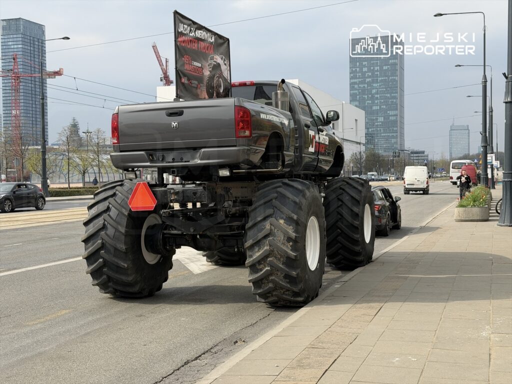 Wysoki monster truck z reklamą "Jazda za kierownicą Monster Trucka" stoi na ulicy obok innych pojazdów.