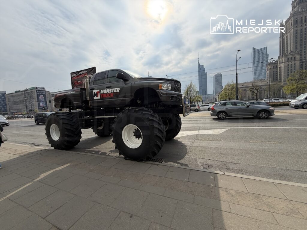 Monster truck z ogromnymi oponami stoi na ulicy, obok ruchu samochodowego i nowoczesnych budynków w tle.