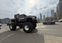 Monster truck z ogromnymi oponami stoi na ulicy, obok ruchu samochodowego i nowoczesnych budynków w tle.