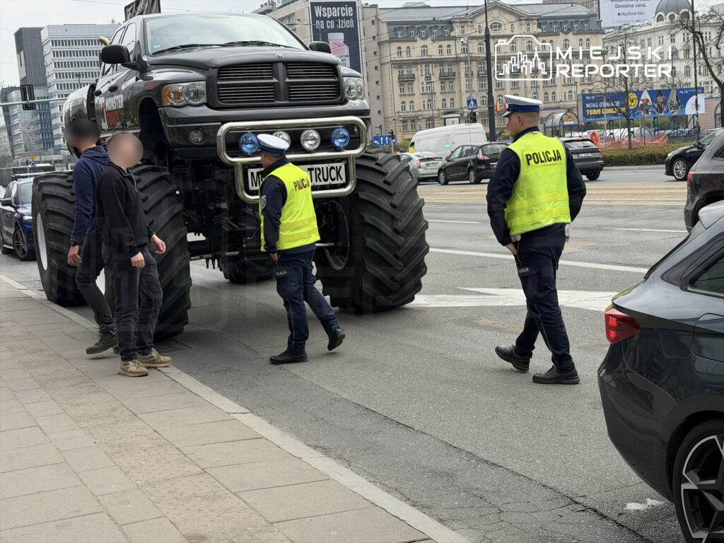 Mężczyzna w mundurze policyjnym rozmawia z dwoma osobami obok dużego samochodu terenowego na ulicy w mieście.