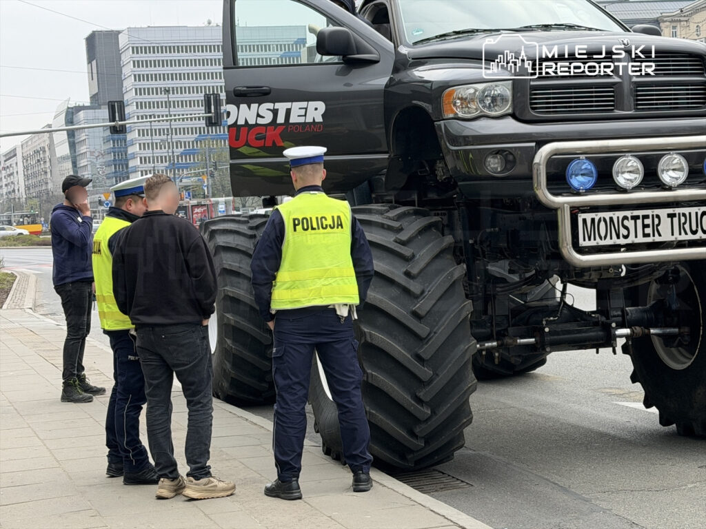 Mężczyźni rozmawiają przy ogromnym monster trucku z napisem "MONSTER TRUCK POLAND" w miejskim otoczeniu.