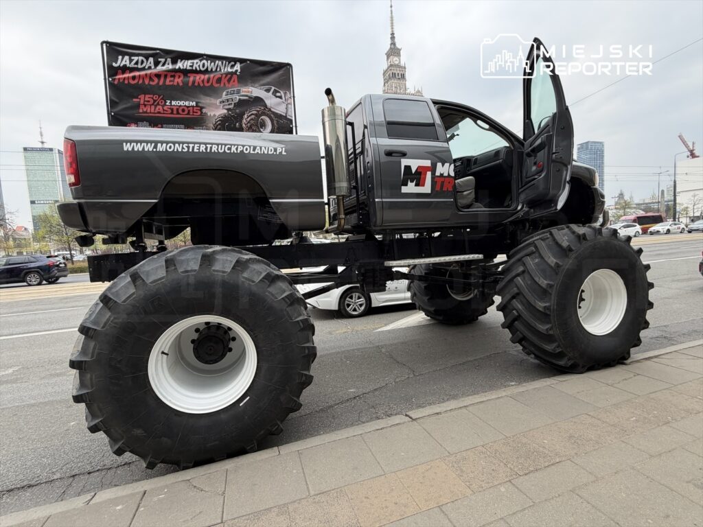 Monster truck z dużymi oponami stoi na ulicy, obok widoczny nowoczesny biurowiec. Na pojeździe reklama jazdy.