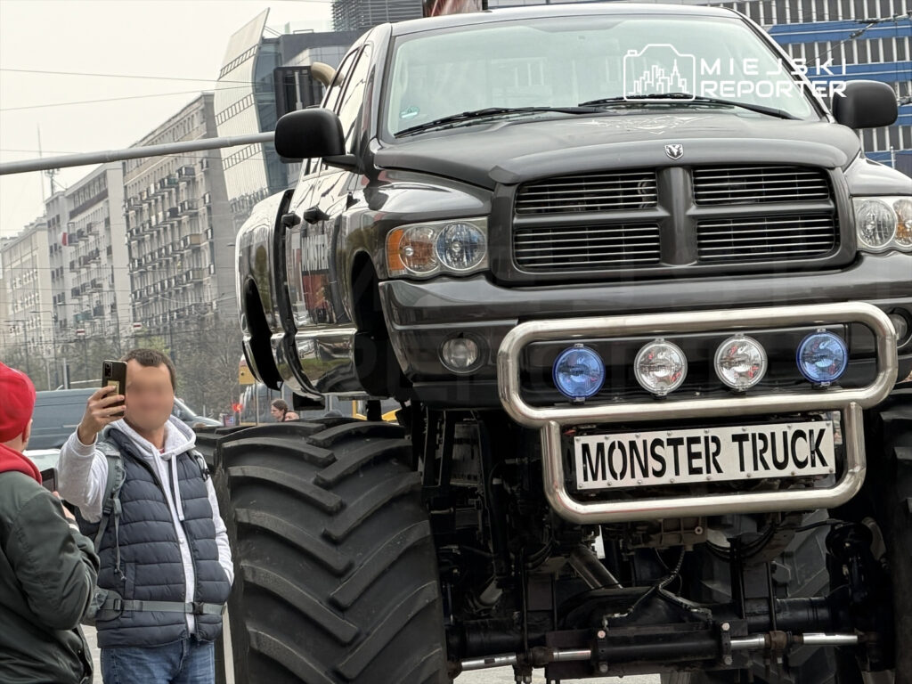Mężczyzna w szarej bluzie robi selfie obok dużego monster trucka z napisem "MONSTER TRUCK" na przednim zderzaku.