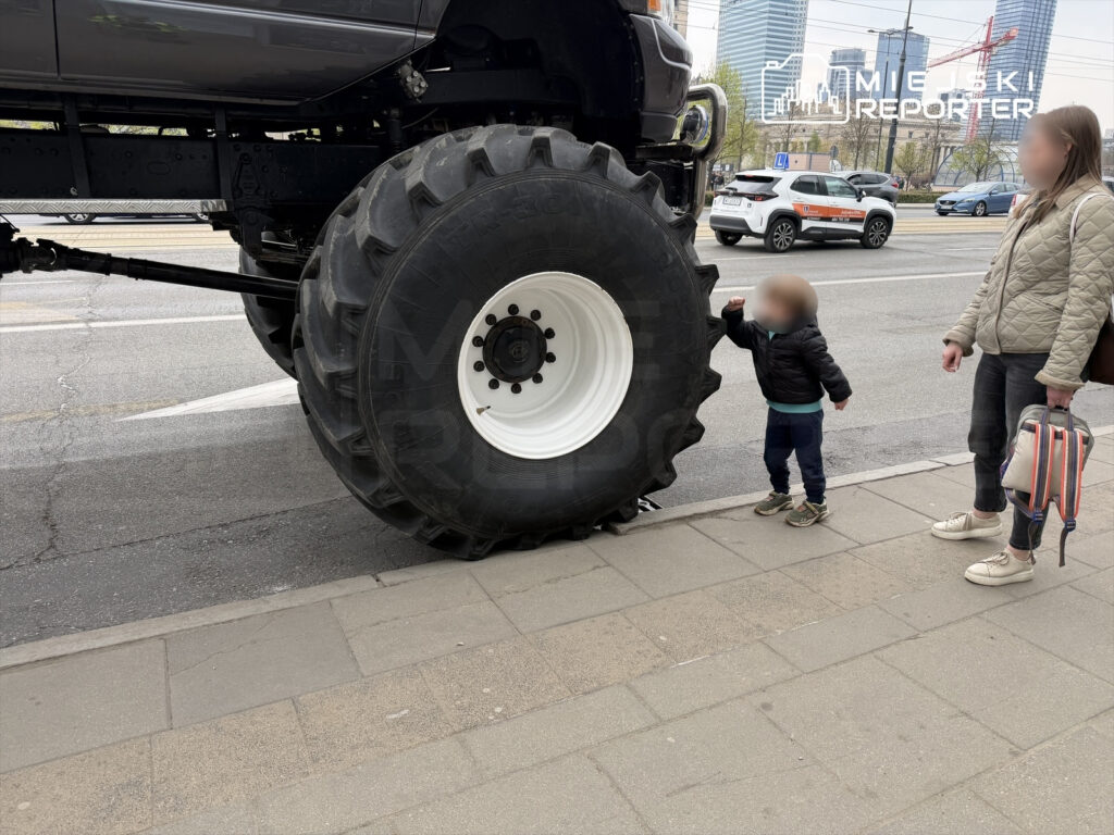 Dziecko w czarnej kurtce dotyka dużej opony pojazdu terenowego, obok stoi kobieta z torbą. W tle widać ruchliwą ulicę.