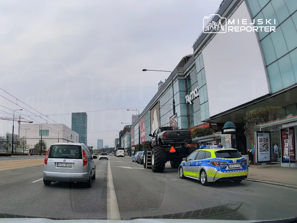 Na ulicy stoi policyjny radiowóz obok dużego samochodu terenowego z ogromnymi oponami, w tle nowoczesny budynek handlowy.