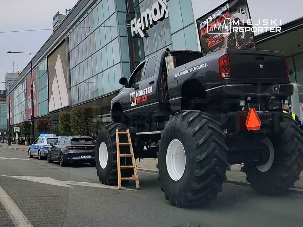 Czarny monster truck z dużymi oponami zaparkowany przy nowoczesnym biurowcu, obok radiowóz policyjny.