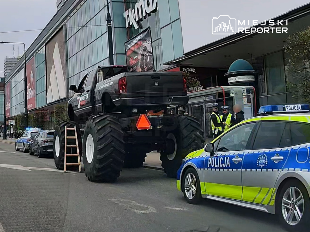 Czarny monster truck z dużymi kołami stoi obok przystanku, obok którego rozmawia dwóch policjantów w kamizelkach odblaskowych.