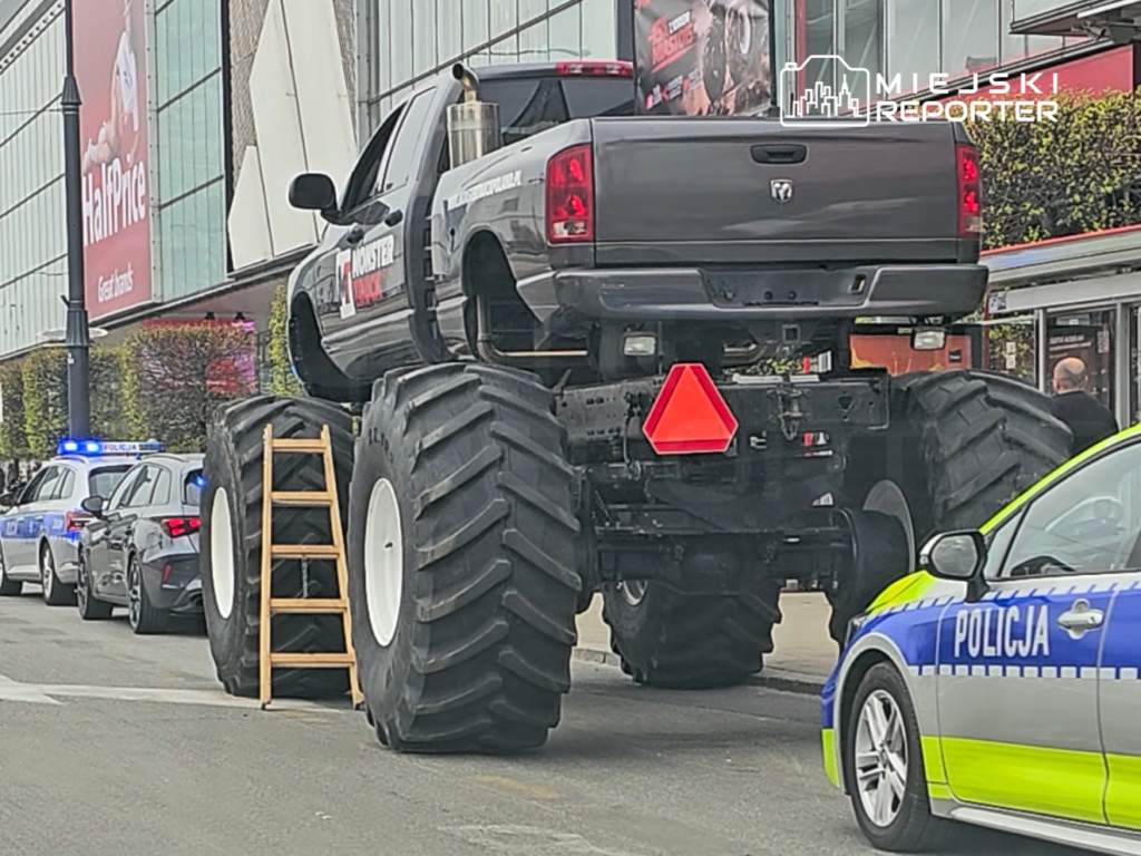 Czarny monster truck z dużymi kołami stoi na parkingu, obok policyjnych radiowozów i drewnianej drabiny.