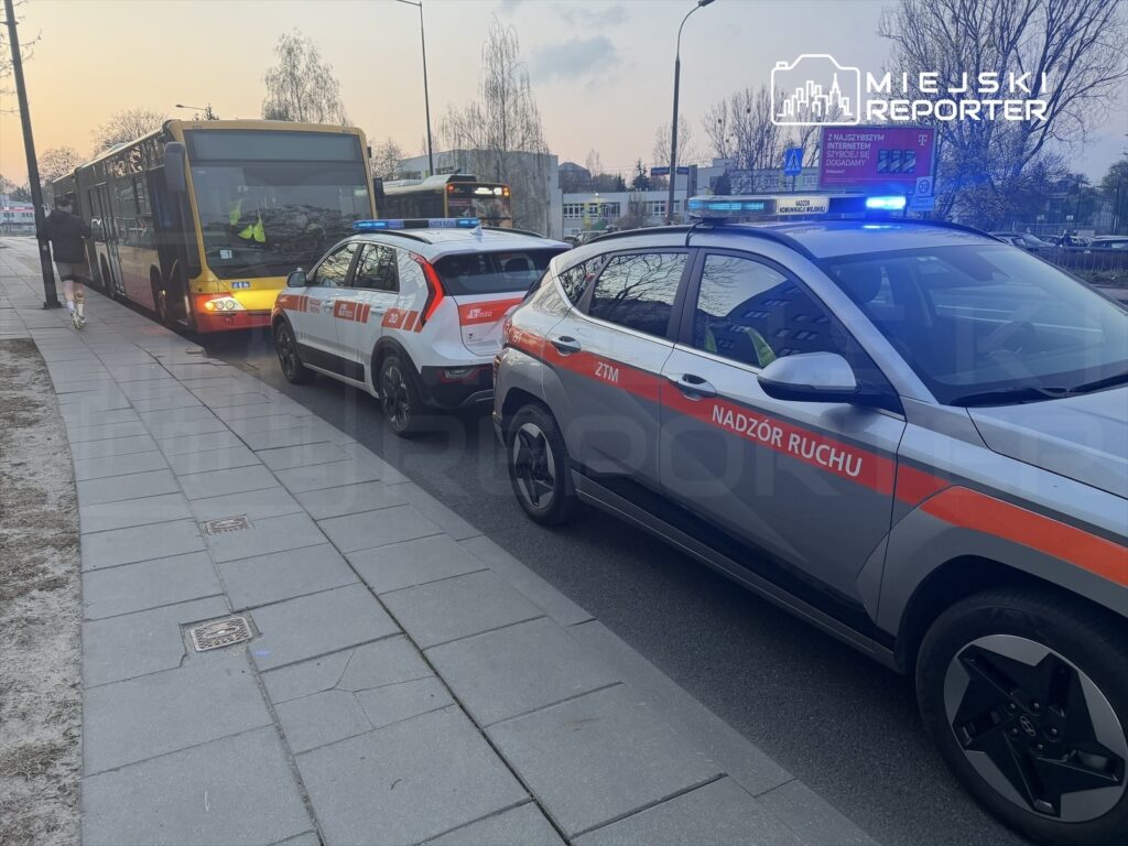 Policyjny radiowóz z włączonymi sygnałami świetlnymi stoi obok przystanku, obok niego kursowy autobus.