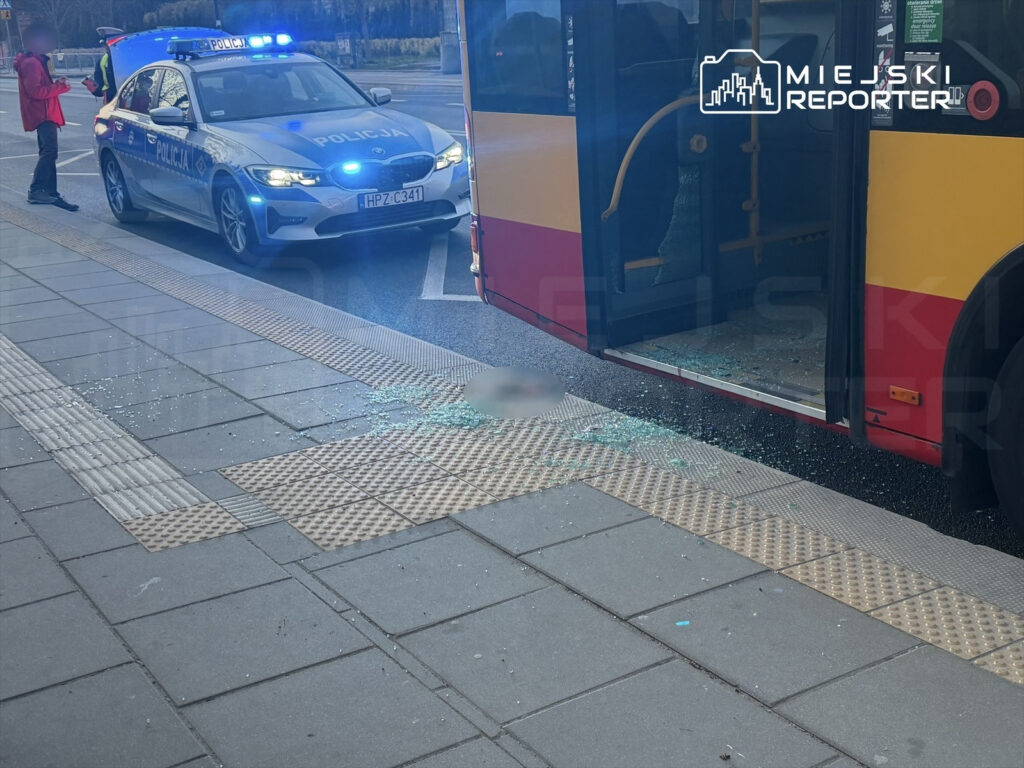 Policjant rozmawia z osobą w czerwonej kurtce obok autobusu miejskiego z rozbitą szybą, na chodniku leżą odłamki szkła.