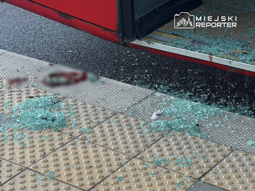 Na chodniku leży rozbite szkło obok otwartych drzwi autobusu, w tle widoczna szara nawierzchnia jezdni.