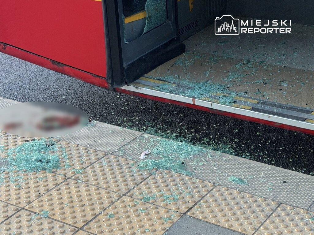 Rozbite szkło leży na chodniku obok otwartych drzwi autobusu miejskiego.