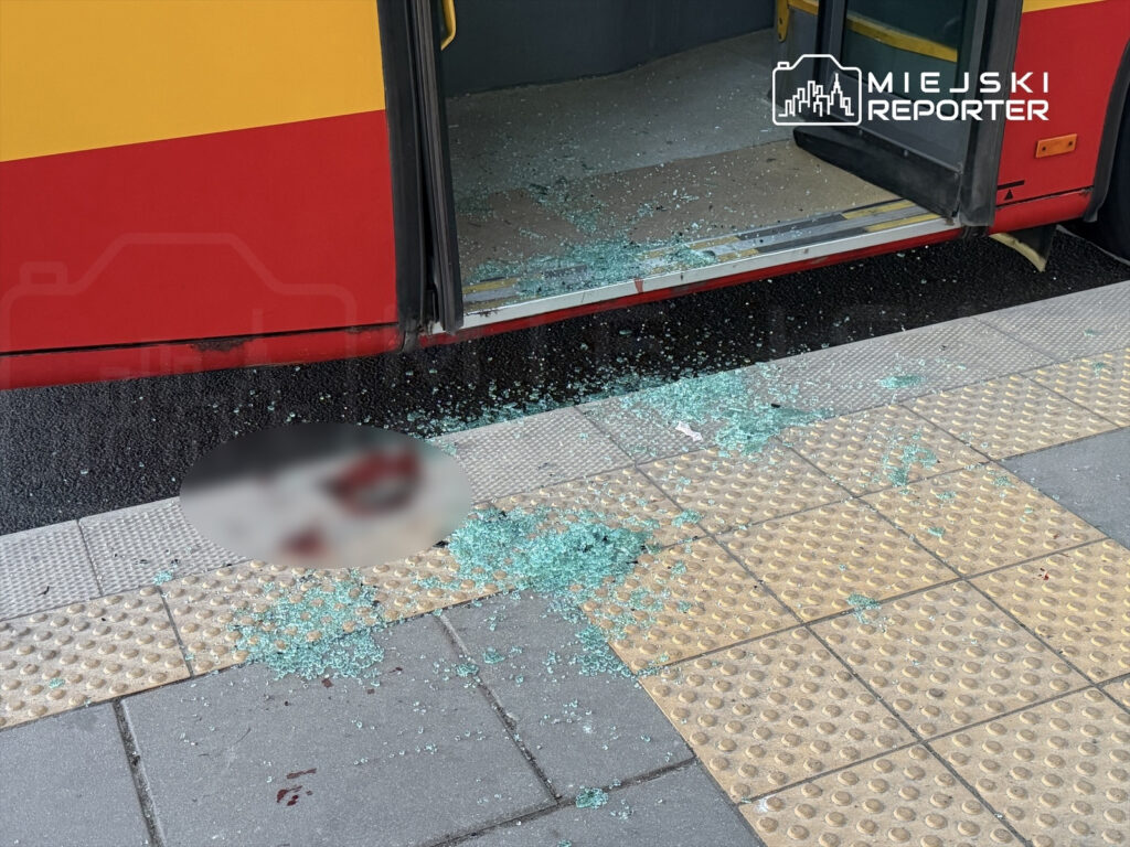 Szkło rozbite na chodniku obok otwartych drzwi autobusu, z plamą na nawierzchni.