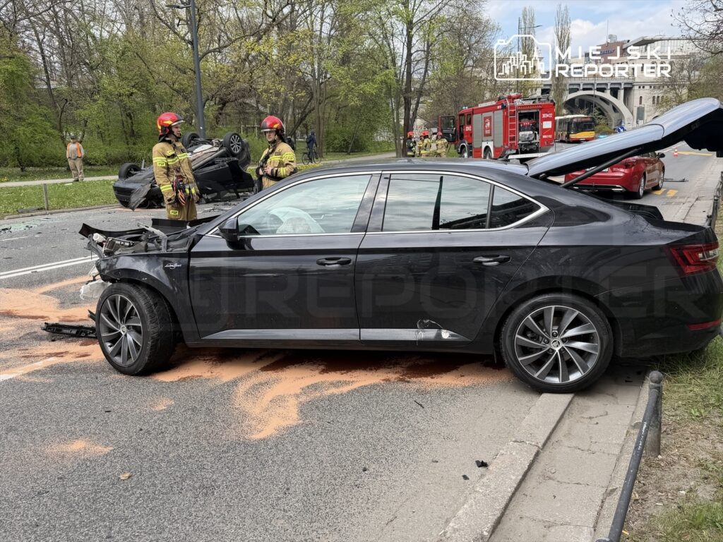 Czarny samochód osobowy stoi na poboczu drogi, obok dwóch strażaków w hełmach, a w tle widoczny odwrócony pojazd i wóz strażacki.