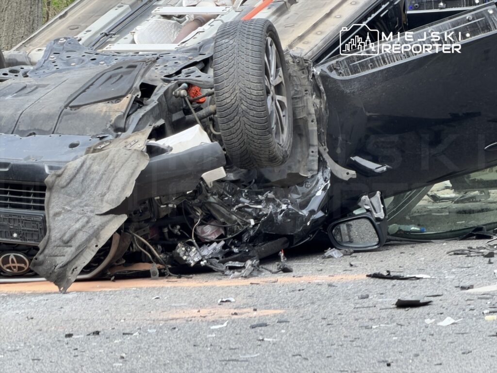 Zniszczony samochód leży na boku na drodze, obok widać odłamki pojazdu i uszkodzone elementy karoserii.