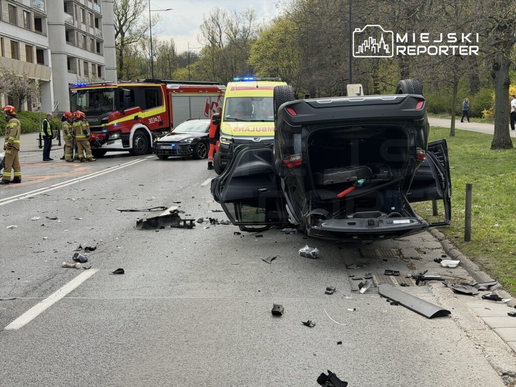 W wyniku wypadku samochodowego przewrócony pojazd leży na drodze, a obok pracują strażacy i służby ratunkowe.