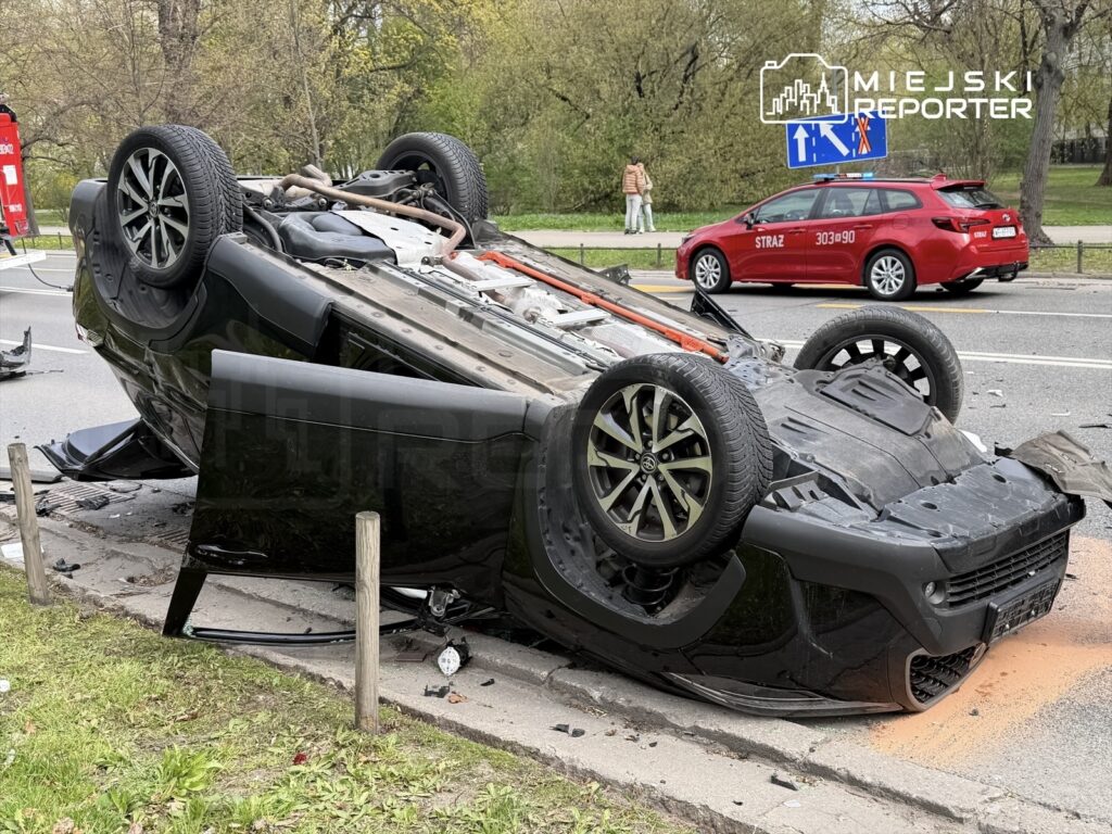 Czarny samochód osobowy leży na dachu na poboczu drogi, obok widoczne fragmenty wraku i straż pożarna w tle.