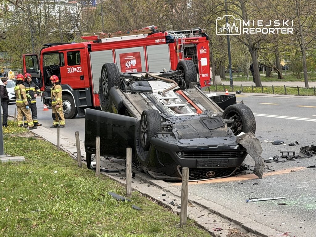 Samochód osobowy przewrócony na dach na jezdni, obok strażacy w mundurach omawiają sytuację przy wozie strażackim.