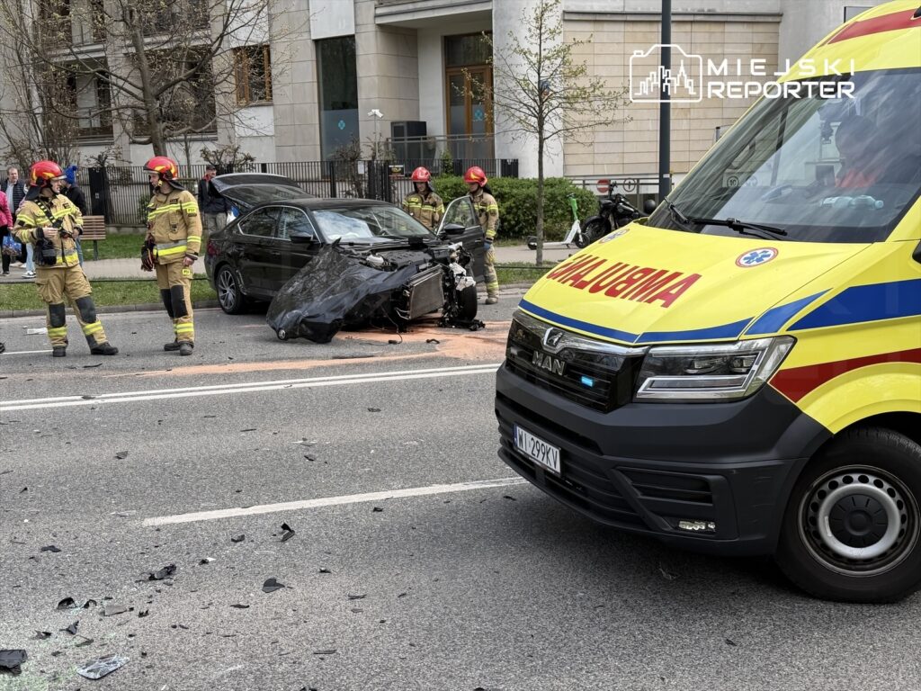 Grupa strażaków w kaskach omawia sytuację obok uszkodzonego samochodu na jezdni, wokół porozrzucane fragmenty pojazdu.