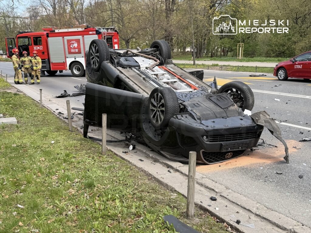 Czarny samochód osobowy leży na dachu obok drogi, a w tle widać strażaków i wóz strażacki.
