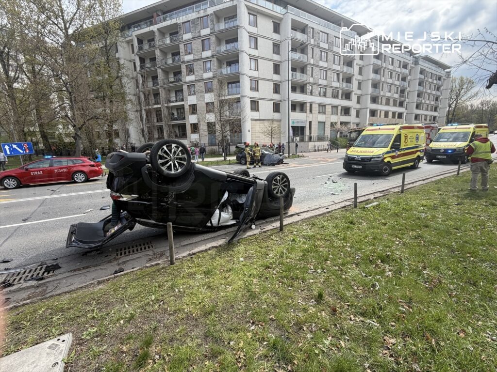 Czarny samochód osobowy leży na dachu na ulicy, obok niego stoją karetki i strażacy, a w tle nowoczesne budynki.