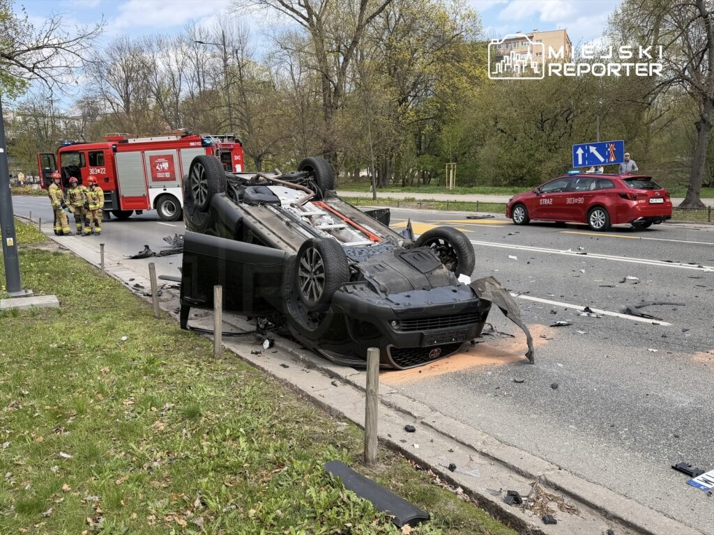 Samochód osobowy leży na dachu na poboczu drogi, obok grupy strażaków oraz wozu strażackiego. W tle widoczna zieleń drzew.