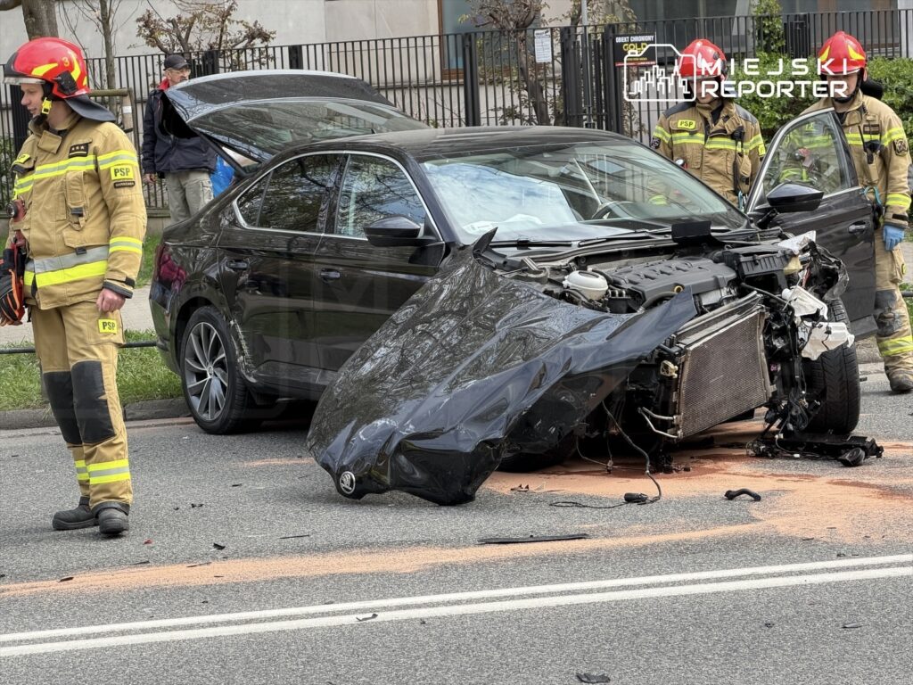 Strażak w mundurze z logo PSP stoi obok uszkodzonego czarnego samochodu na drodze, wokół widoczne ślady wypadku.