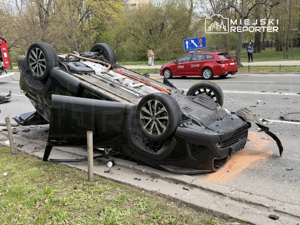Czarny samochód osobowy leży na dachu na jezdni, obok widoczna straż pożarna i oznakowanie drogowe.