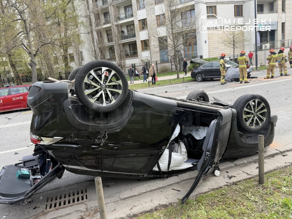Czarny samochód osobowy leży na dachu na ulicy, obok pracują strażacy oraz zniszczone inne pojazdy.