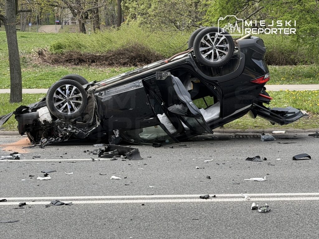 Czarny samochód osobowy leży na dachu na jezdni, wokół rozrzucone fragmenty karoserii i inne zniszczenia.