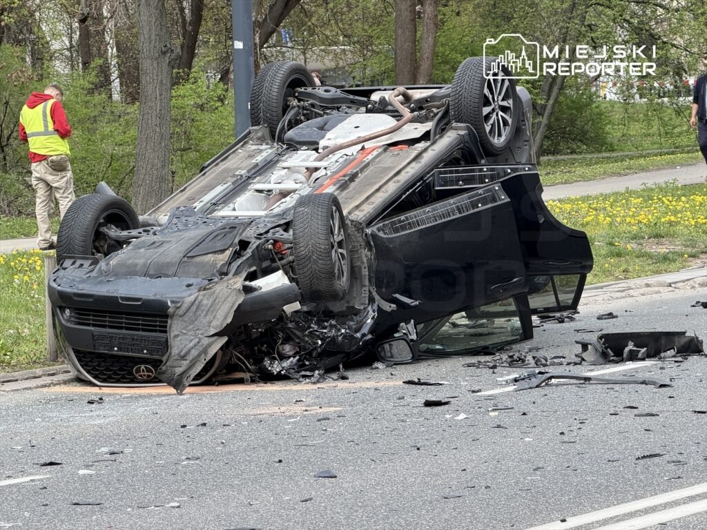 Samochód leżący na dachu na jezdni, wokół rozrzucone fragmenty pojazdu, w tle mężczyzna w żółtej kamizelce.