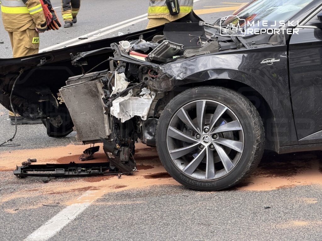Uszkodzony samochód osobowy z rozbitym przodem i wyciekającym płynem, na drodze obok strażaków w mundurach.