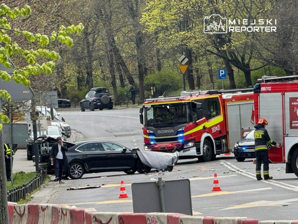 Czarny samochód osobowy stoi na drodze, obok interweniują strażacy i pojazdy ratunkowe, a na jezdni leżą fragmenty rozbitego pojazdu.