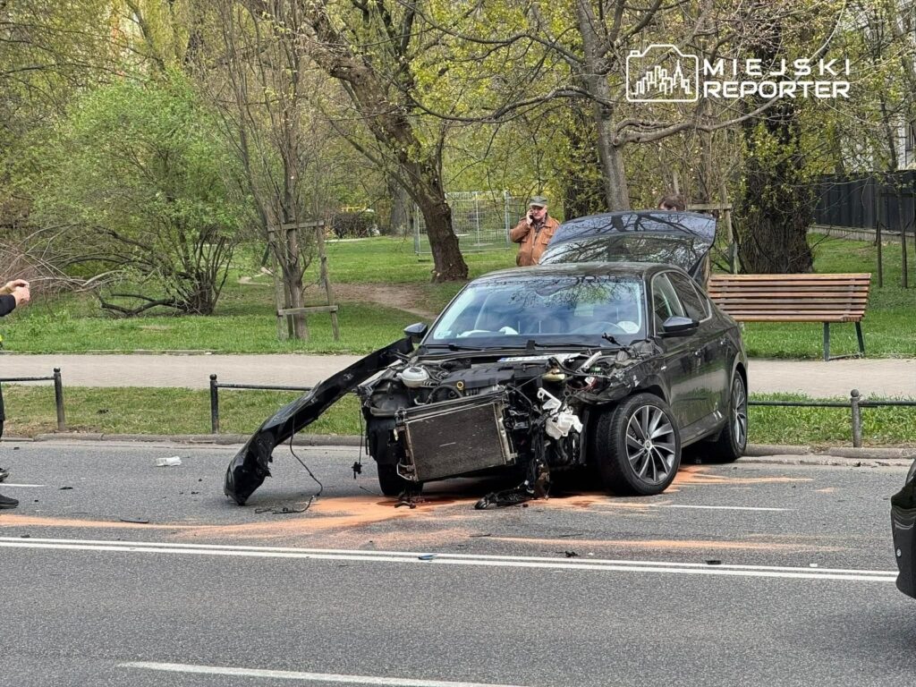 Czarny samochód osobowy z uszkodzoną przednią częścią stoi na jezdni, obok mężczyzna robi zdjęcia. W tle park z ławką.