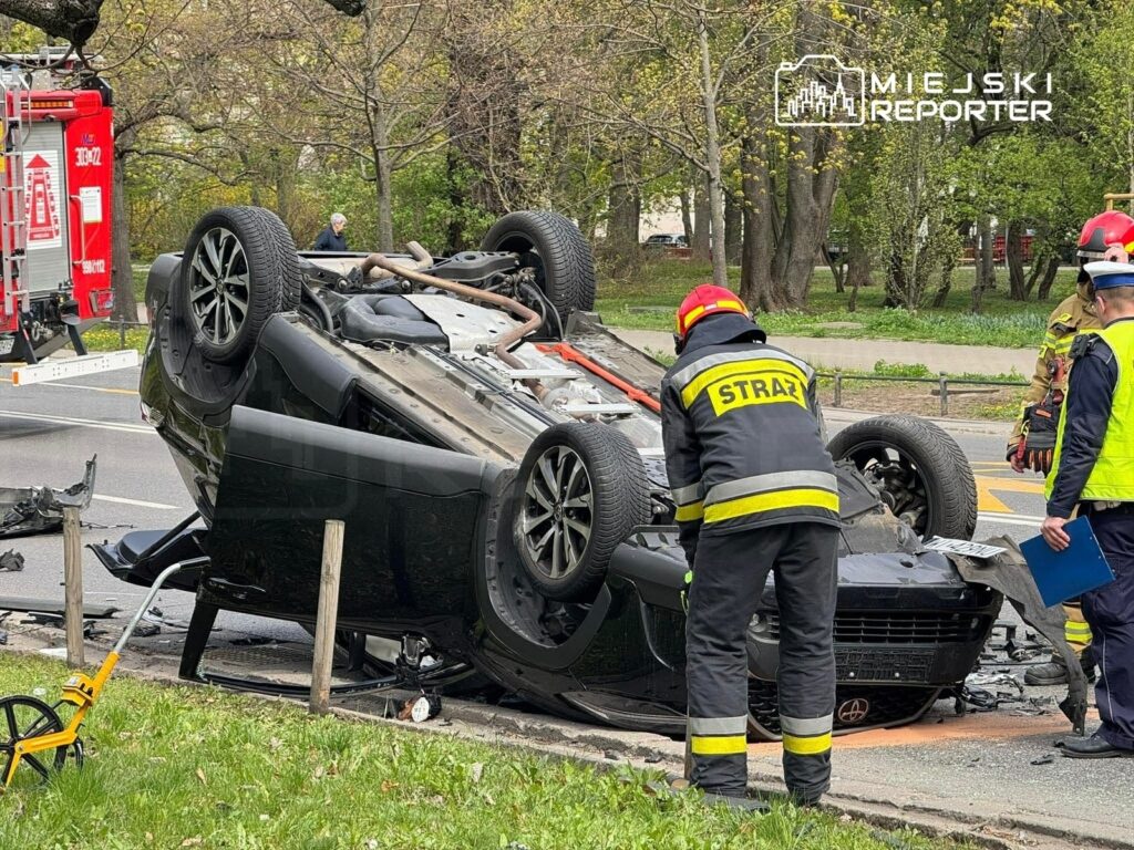 Strażacy w mundurach pracują przy przewróconym samochodzie osobowym na drodze, obok widoczny wóz strażacki.