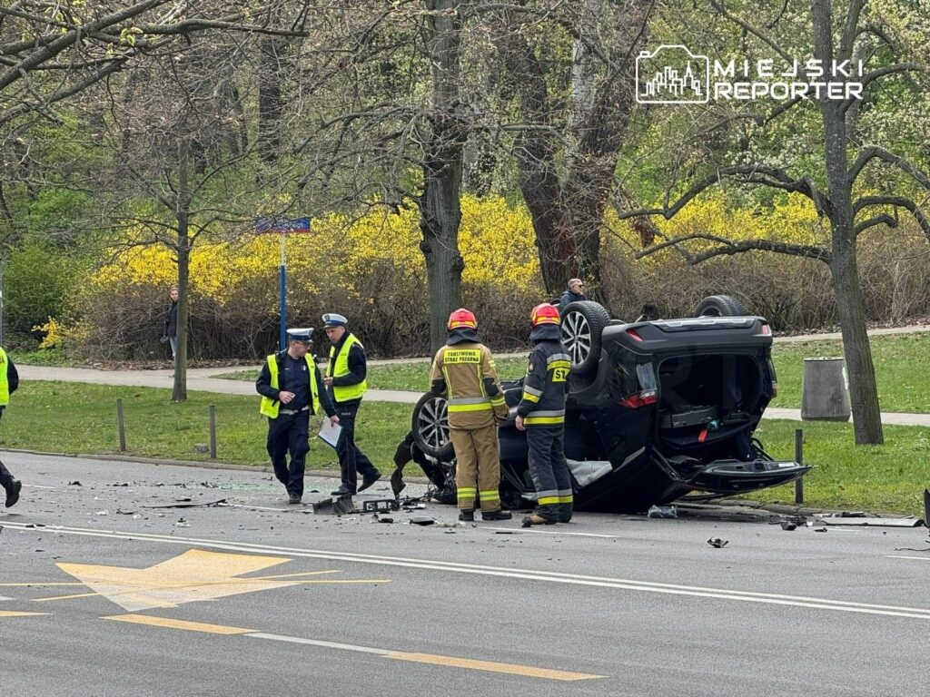 Na drodze leży przewrócone auto, a obok pracują strażacy i policjanci, zbierając odłamki szkła. W tle widać zieleń i kwitnące krzewy.