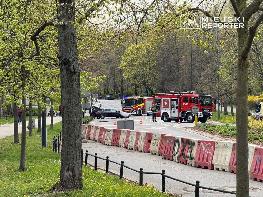 W tle widać straż pożarną interweniującą przy wypadku drogowym na ulicy, z ustawionymi pachołkami.