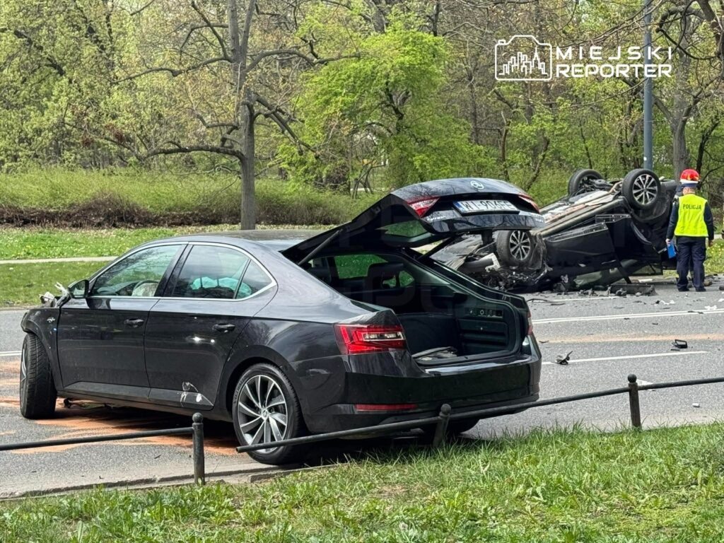 Na drodze leży przewrócone auto osobowe, a obok stoi policyjny funkcjonariusz z notatnikiem. W tle widoczna jest uszkodzona limuzyna.