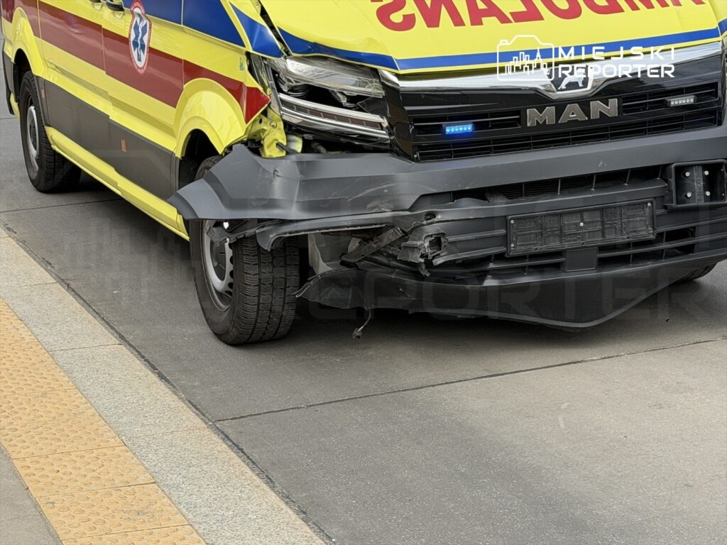 Uszkodzony ambulans z wgniecionym przodem stoi na miejskiej ulicy w pobliżu chodnika.