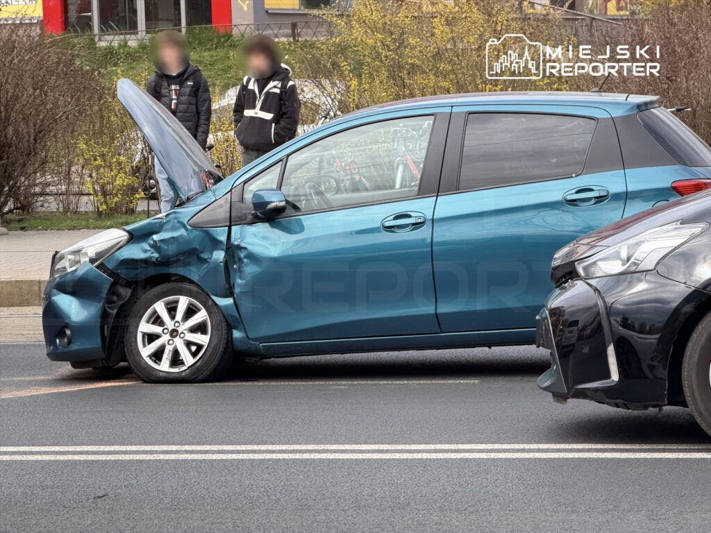 Zniszczony niebieski samochód osobowy z otwartą maską stoi na drodze, obok dwóch osób obserwujących sytuację.