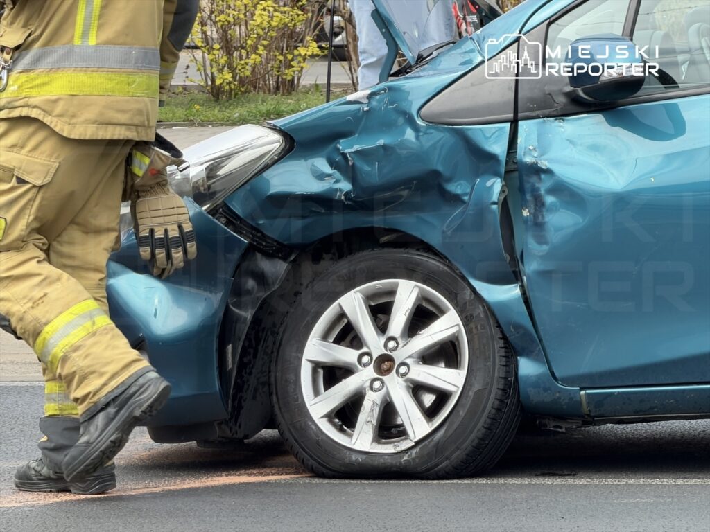 Strażak w mundurze zbliża się do uszkodzonego, niebieskiego samochodu osobowego na drodze.