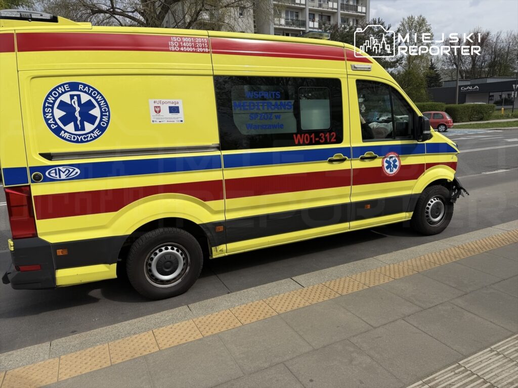 Żółta karetka pogotowia z logo Państwowego Ratownictwa Medycznego zaparkowana na chodniku w miejskim otoczeniu.