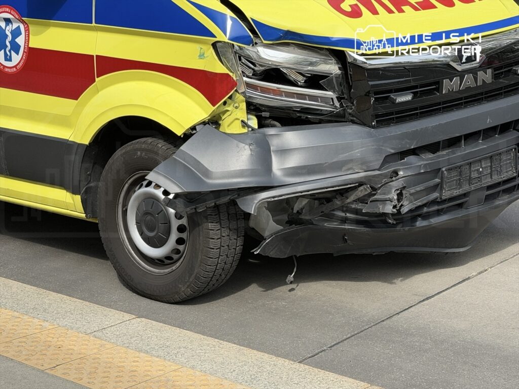 Uszkodzony ambulans z wgniecionym przodem zaparkowany na chodniku w miejskim otoczeniu.